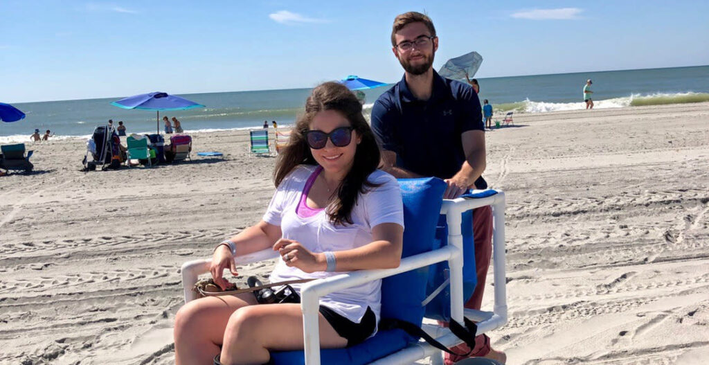 brunette woman wearing sunglasses, white and pink t-shirt sitting in blue cushioned beach wheelchair being pushed by man with glasses wearing a blue shirt. ocean and beach in background