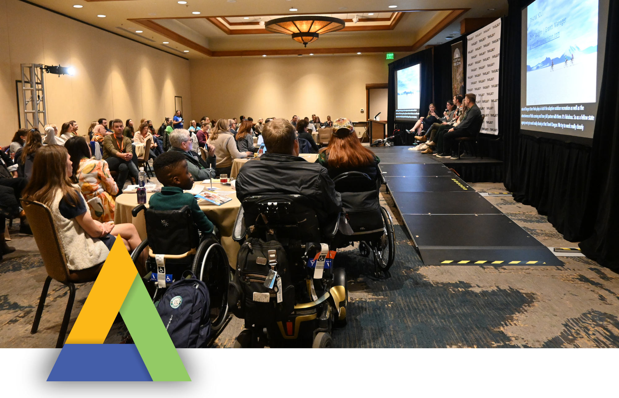 A large conference room filled with attendees seated in rows and at round tables, listening to a panel discussion on stage. Several attendees use wheelchairs, and the panelists sit in a row at the front beside a large screen displaying slides with captions. A long black ramp leads to the stage, and the TravelAbility logo sits overlaid in the bottom left corner of the image. The atmosphere appears engaged and attentive.
