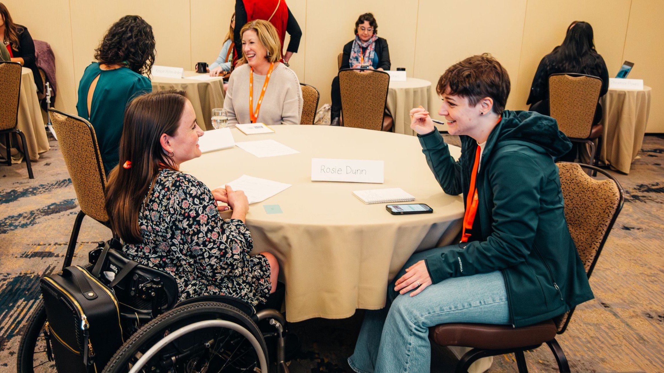 Small group discussion at a conference, with several adults seated around round tables in a warm, collaborative setting. In the foreground, two people talk and smile across a table, one using a wheelchair, with name cards and notebooks visible.