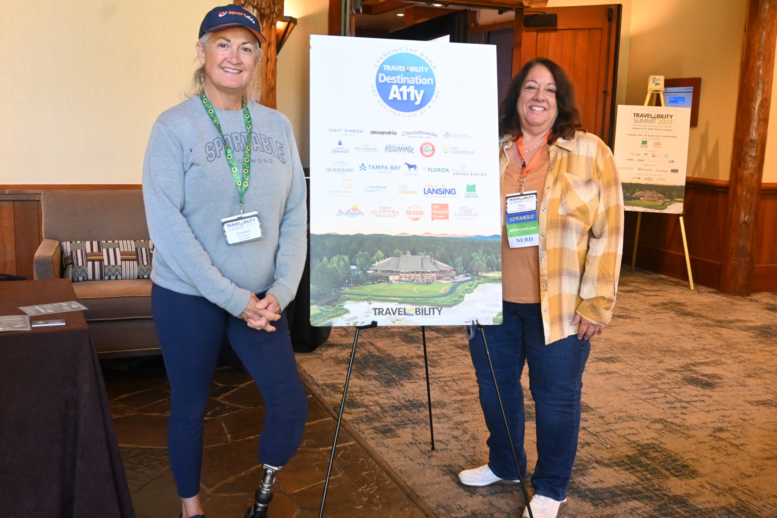 Toni Bastian of Visit Richmond and her guest, Suellen Henneberry, stand in front of a Destination A11y Club sign at the 2025 TravelAbility Summit. Toni wears a plaid shirt and jeans, while Suellen wears a gray sweatshirt, dark pants, and a baseball cap and has a prosthetic leg.